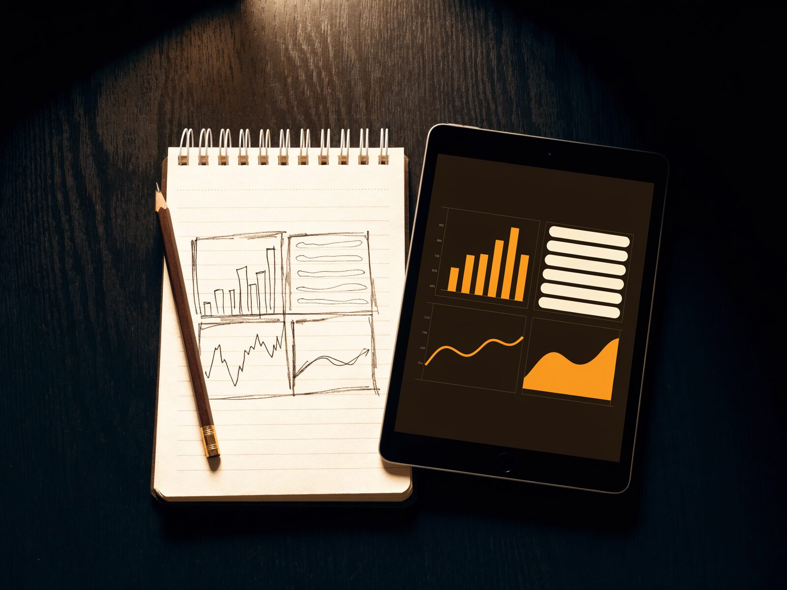 A pencil sketch of a four-panel dashboard in a notepad beside a tablet showing the same dashboard finished, on a dark wooden desk under warm lamp light